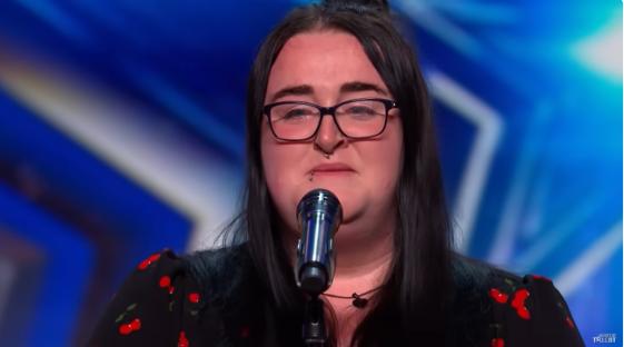 A woman with glasses and a cherry-printed shirt cries while speaking into a microphone.