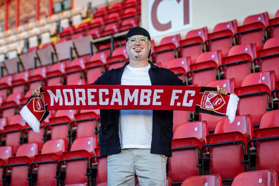Man holding Morecambe FC scarf in stadium seats.