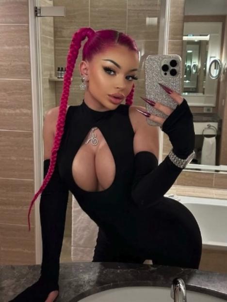 A woman with pink pigtail braids, in a black cutout jumpsuit, holding a phone for a mirror selfie.
