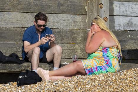 Gemma Collins and a friend at the beach; she's smoking and he's on his phone.