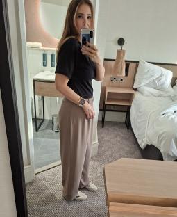 Woman taking a selfie in a hotel room, wearing taupe pants and a black shirt.