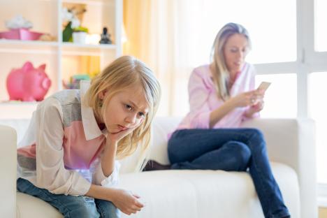 Sad young girl sitting on a couch while her mother texts on her phone.