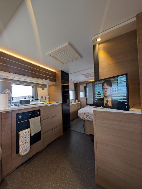 Interior view of a caravan with a kitchen and bedroom area, showing a TV playing *The Office*.