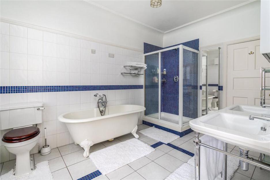 A bathroom with a white clawfoot tub, a tiled shower with blue mosaic tiles, a toilet with a dark wooden seat, and two pedestal sinks.