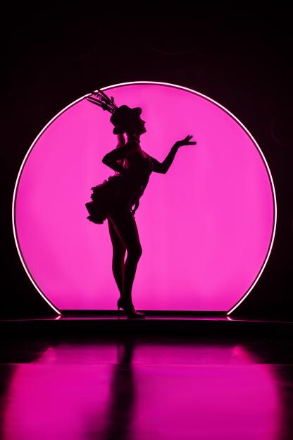 Silhouette of a performer in a feathered hat and ruffled outfit standing in front of a large pink-lit circle.
