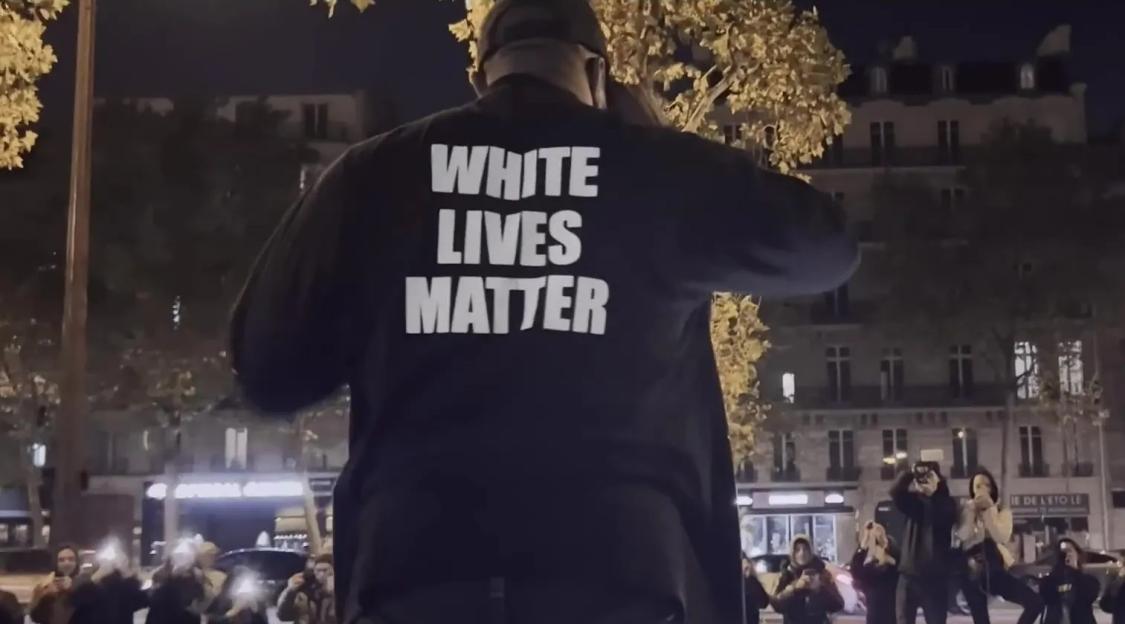 A man wearing a "WHITE LIVES MATTER" shirt speaking to a crowd at night.