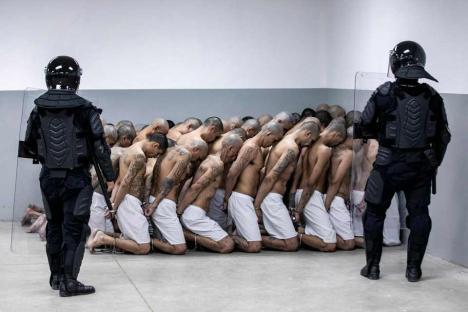 Handcuffed and kneeling tattooed prisoners flanked by riot police.