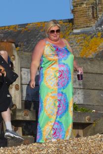 Gemma Collins on a beach, wearing a patterned maxi dress and sunglasses, holding a drink.