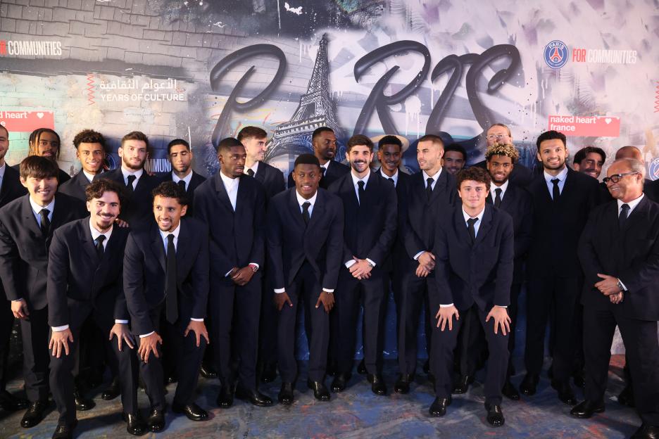 A group of men in dark suits and black ties pose for a photo in front of a graffiti-style mural of "Paris" and the Eiffel Tower.