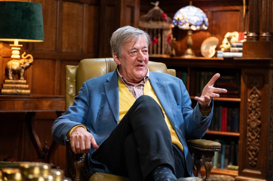 Stephen Fry sits in a leather armchair in a wood-paneled room.