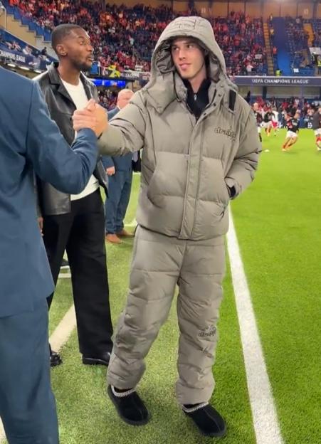 A young man in a padded gray jacket and matching pants, with a hood pulled over his head, shakes hands with another person on a football field.