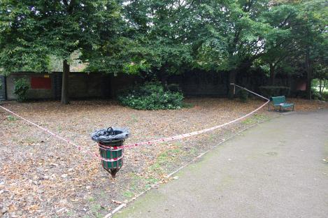 Crime scene in a park with police tape surrounding a trash can and a park bench.
