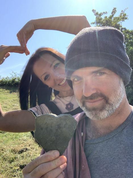 A couple posing for a photo; the woman makes a heart shape with her hands above the man's head, while he holds a heart-shaped rock.