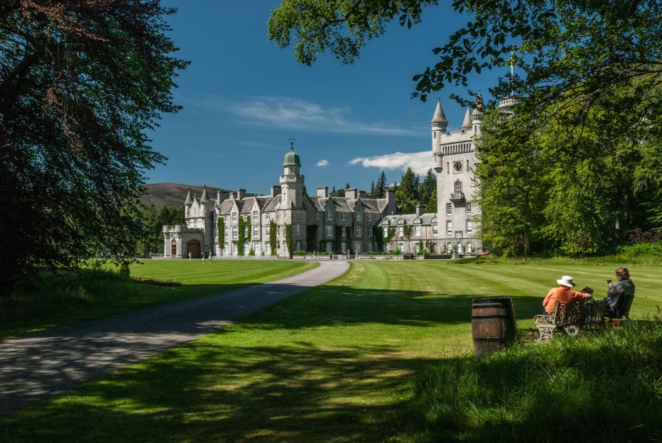 Balmoral Castle in Aberdeenshire, seen across a large green lawn with two people relaxing on a bench in the foreground.