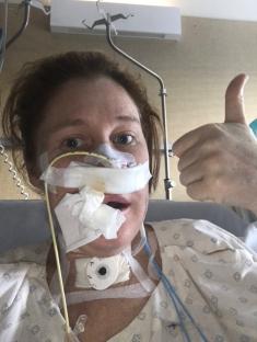 Woman in hospital bed giving thumbs up, post-facial surgery.