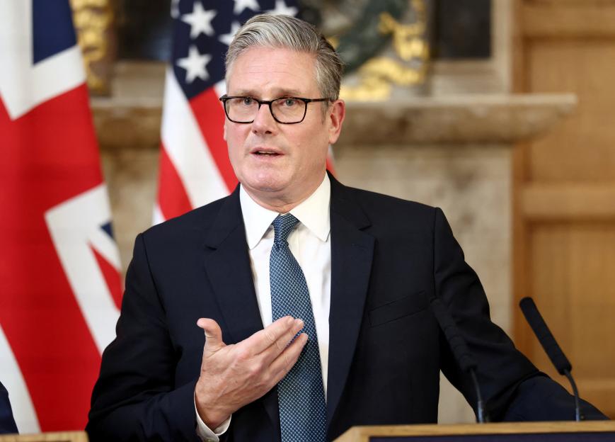 British Prime Minister Keir Starmer speaking at a press conference with Union Jack and American flags in the background.