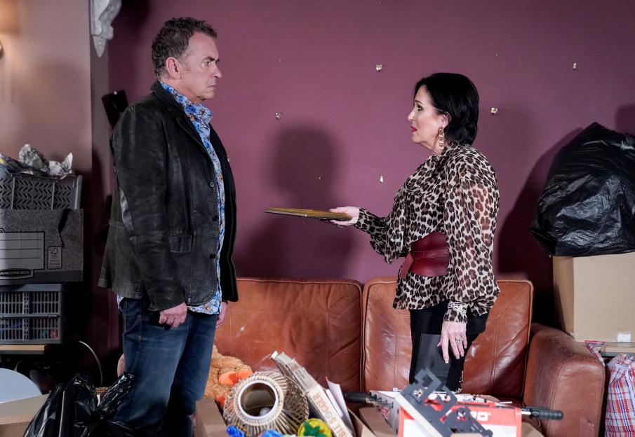 A woman hands a man a book in a cluttered room.
