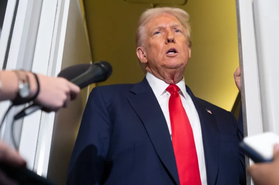 Donald Trump in a dark suit and red tie, speaking to microphones.