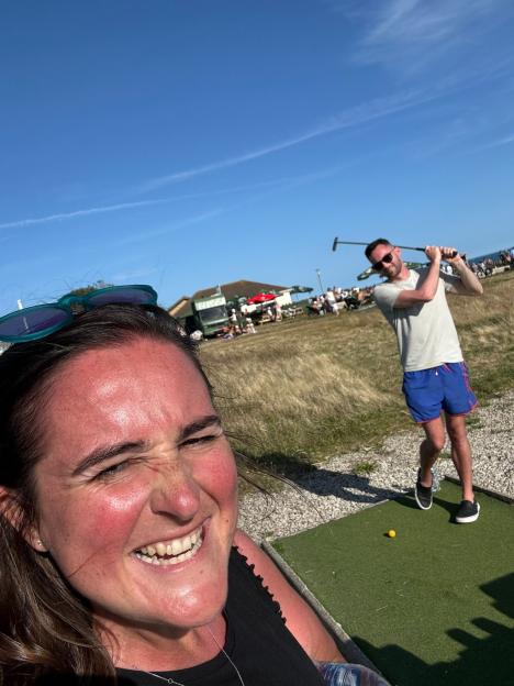 Woman smiling in foreground with man playing golf in background.