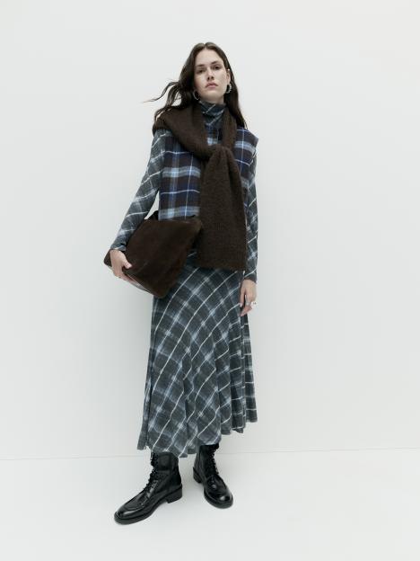 A model wearing a grey plaid long-sleeved dress, a blue and brown plaid vest, a brown knitted scarf, black boots, and carrying a brown bag.
