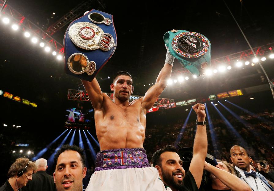 Amir Khan celebrates a boxing victory, holding two championship belts.