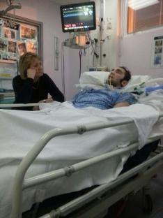 Woman sitting beside a man lying in a hospital bed.