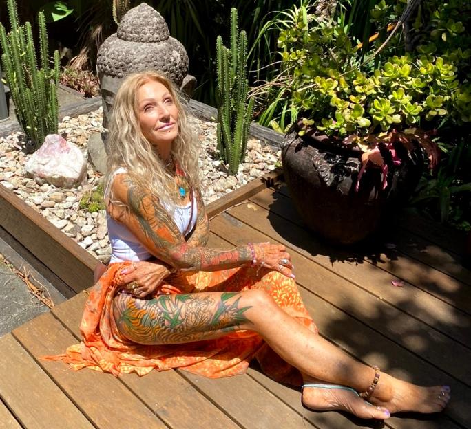 Woman with long blonde hair and colorful tattoos on her arms and legs sitting on a wooden deck.