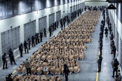 Hundreds of inmates sitting on the floor of a prison, surrounded by guards.
