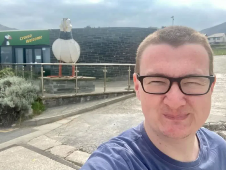 Rich James Cross, 27, in a blue shirt and glasses, smiling for a selfie in front of a building with a "Croeso Welcome" sign and a large bird statue.
