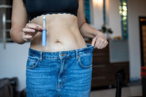 A woman celebrates her weight-loss progress by holding a semaglutide injector in one hand and pulling on the waistband of jeans that no longer fit, showing her transformation.