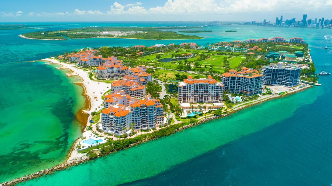 Aerial view of Fisher Island, Miami, Florida, showing residential buildings, green spaces, and beaches surrounded by turquoise water.