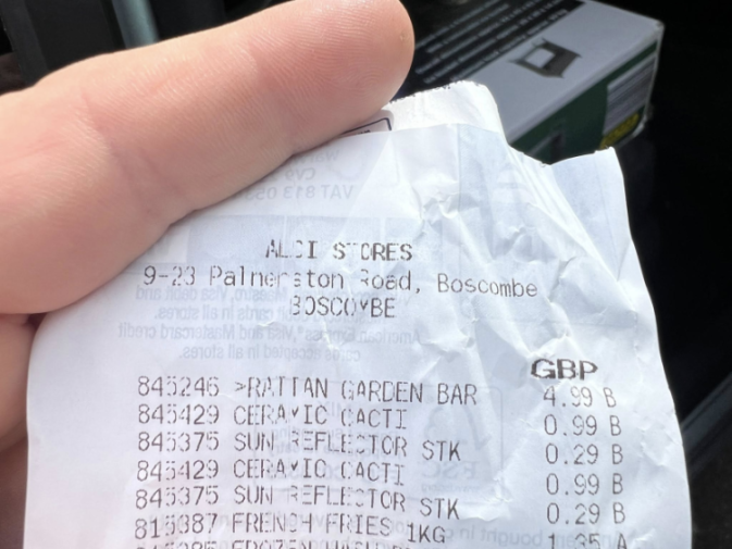 A person holding an Aldi receipt showing the purchase of a rattan garden bar for £4.99, ceramic cacti for £0.99, and sun reflector stakes for £0.29.