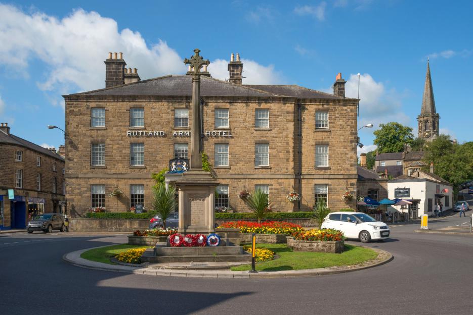 Rutland Arms Hotel in Bakewell, Derbyshire, with a war memorial in the foreground surrounded by flowers.
