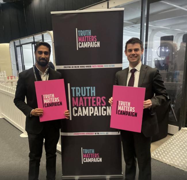 Two men hold pink "Truth Matters Campaign" signs next to a campaign banner.