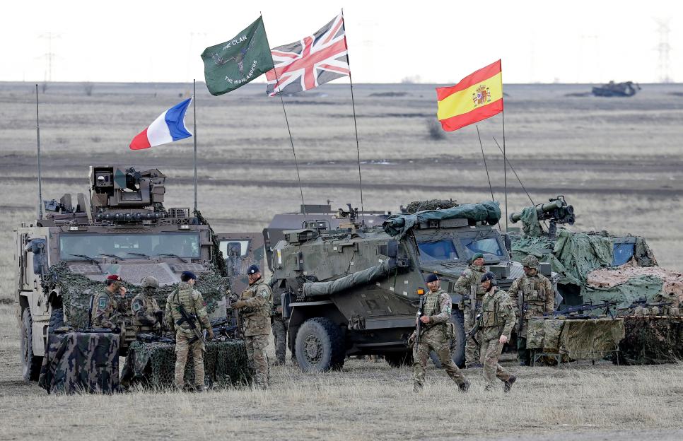 Soldiers stand next to armored vehicles, with flags from France, the UK, and Spain waving in the background, during the NATO Exercise Steadfast Dart 25.