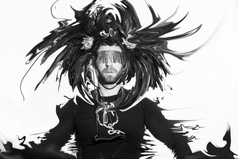 Black and white illustration of a man with a beard wearing a metal blindfold and necklaces, surrounded by swirling abstract shapes.