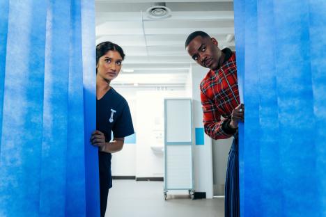 A doctor and a nurse peeking from behind blue curtains in a hospital hallway.