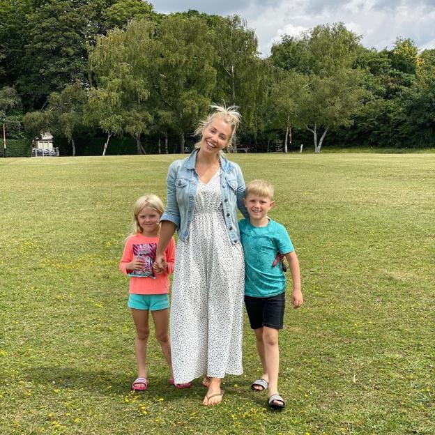 A smiling woman holding hands with her two children in a grassy park.