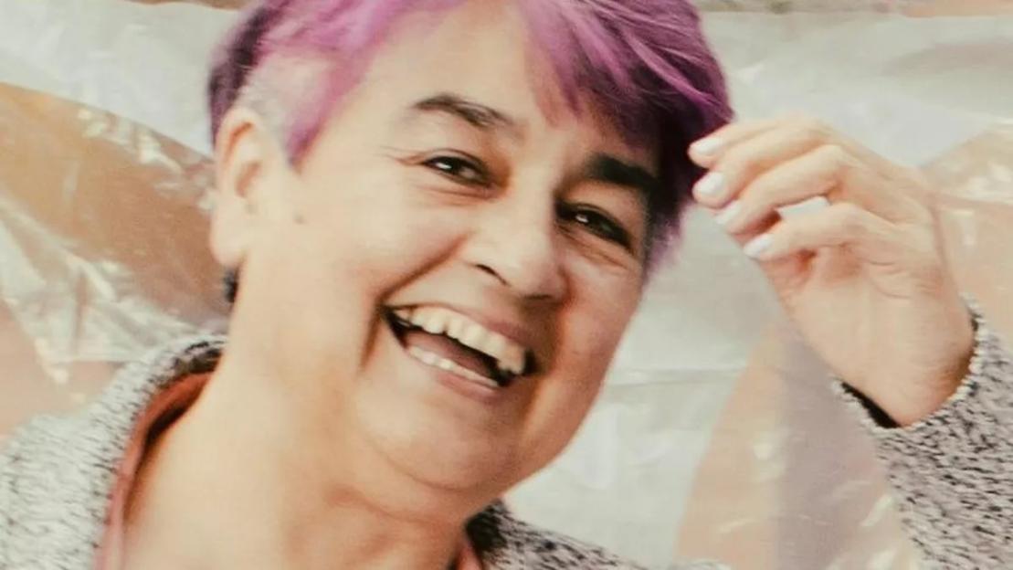 Bernadette Rosario, a woman with short purple hair, smiling and holding her hand to her hair.
