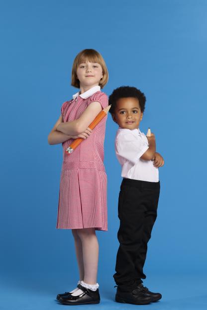 Two children in school uniforms stand back-to-back holding large pencils.
