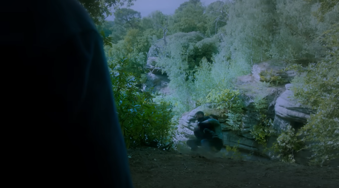 Screenshot of two people struggling near a rocky outcrop in a wooded area.