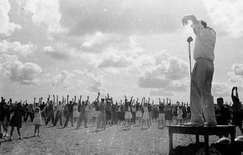 Black and white photo of a large group of people participating in a mass exercise session led by an instructor.