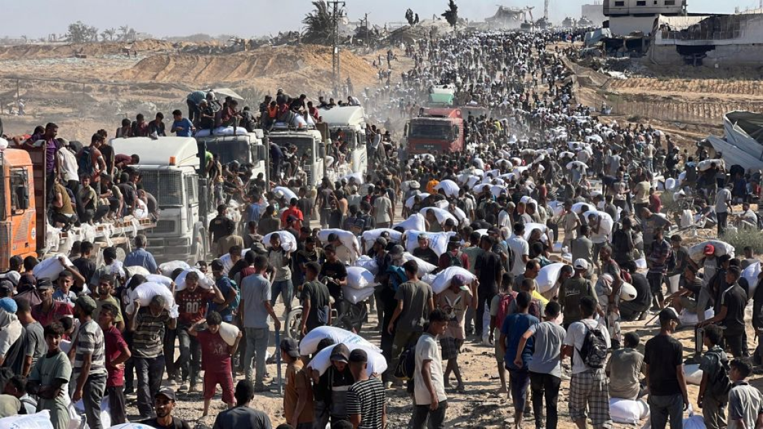 Large crowd of people carrying sacks, many crowded onto trucks.