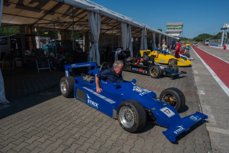 A man sits in a blue Formula One race car in a pit lane.