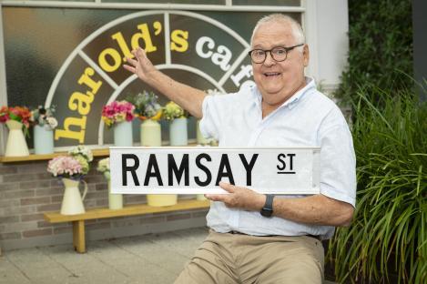 Ian Smith holding a Ramsay Street sign in front of Harold's Cafe.