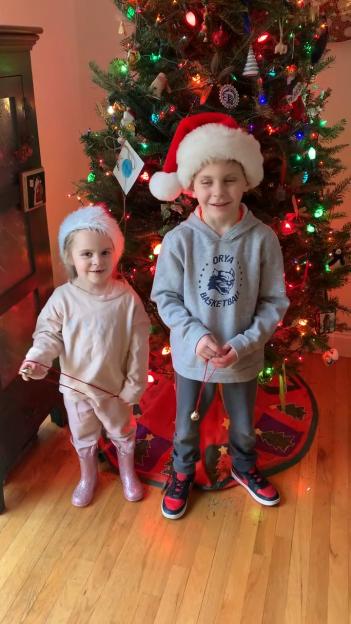 Two children in Santa hats stand in front of a Christmas tree.