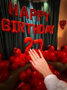 A hand displaying a diamond ring in front of a 27th birthday balloon display.