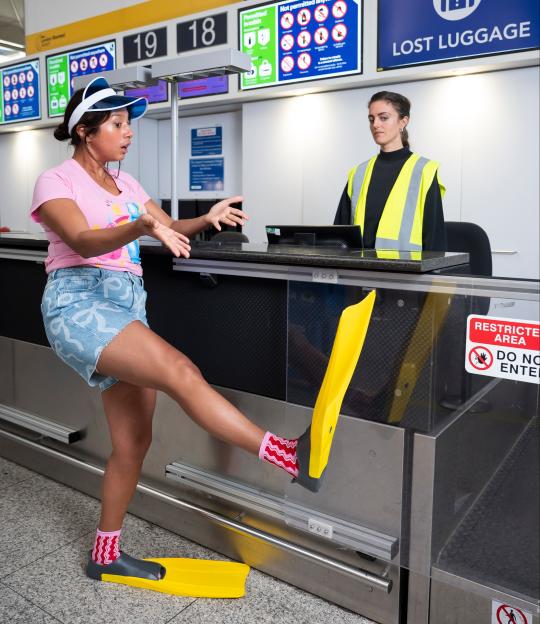 A traveler with flippers on her feet and pink socks discusses a lost item with an airport employee at the check-in desk, highlighting the importance of travel insurance.