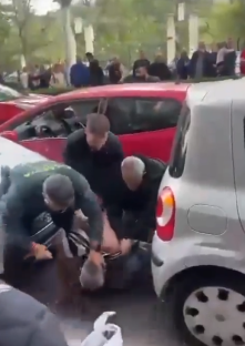 People restraining a person on the ground near damaged cars.