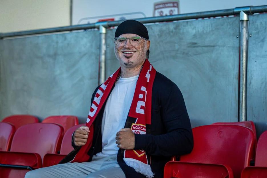 Man wearing a Morecambe FC scarf smiles at the camera.